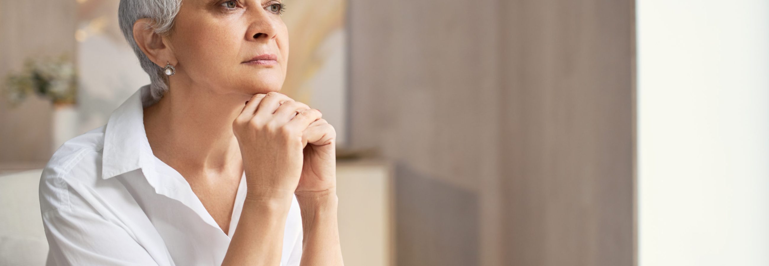 Serious elegant retired woman with short hairstyle posing indoors with hands under chin, looking away with pensive facial expression, thinking over some idea or decision, being deep in thoughts