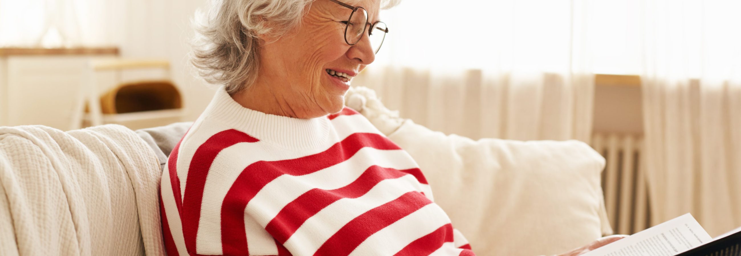 Side view of cute happy grandmother in glasses enjoying reading indoors, sitting on sofa with interesting detective story, smiling joyfully. Stylish elderly woman relaxing on couch holding book